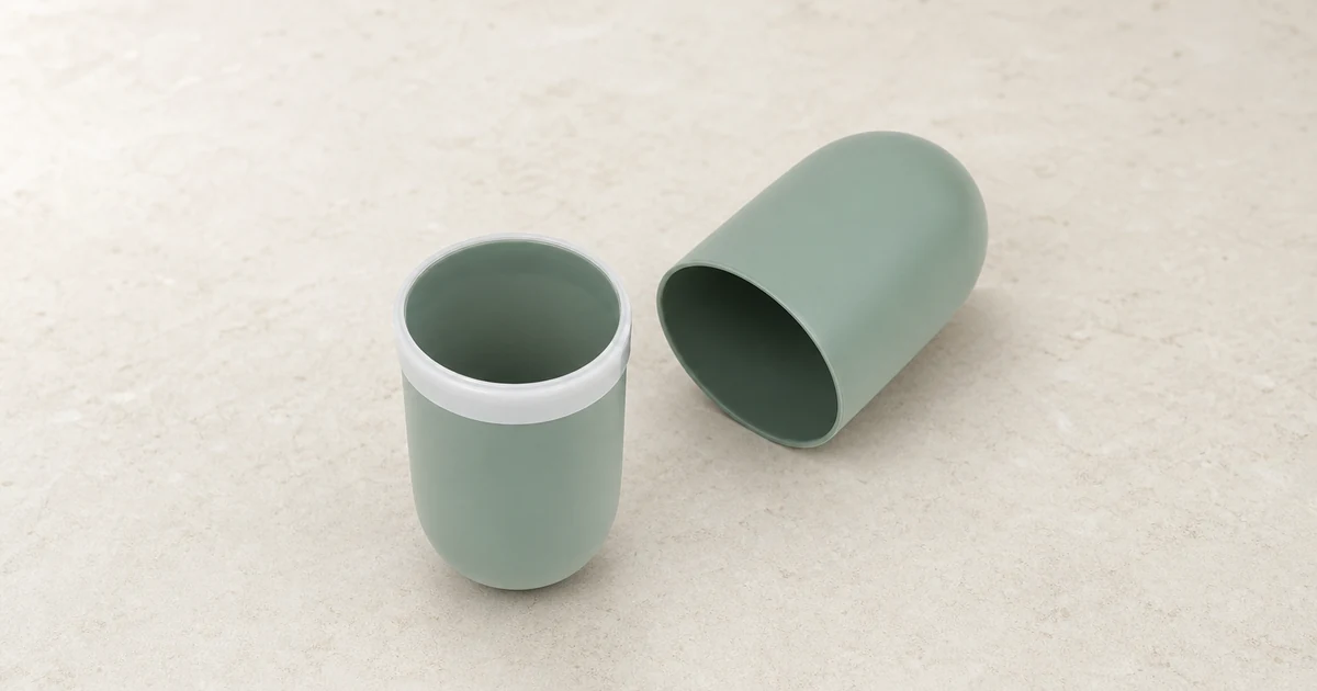 Un estuche de viaje verde salvia para cepillo, abierto en dos mitades con forma de vaso sobre una encimera de baño